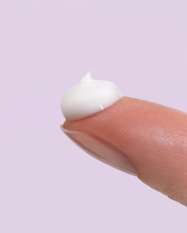 Pequena quantidade de creme branco na ponta de um dedo, sobre fundo lilás.