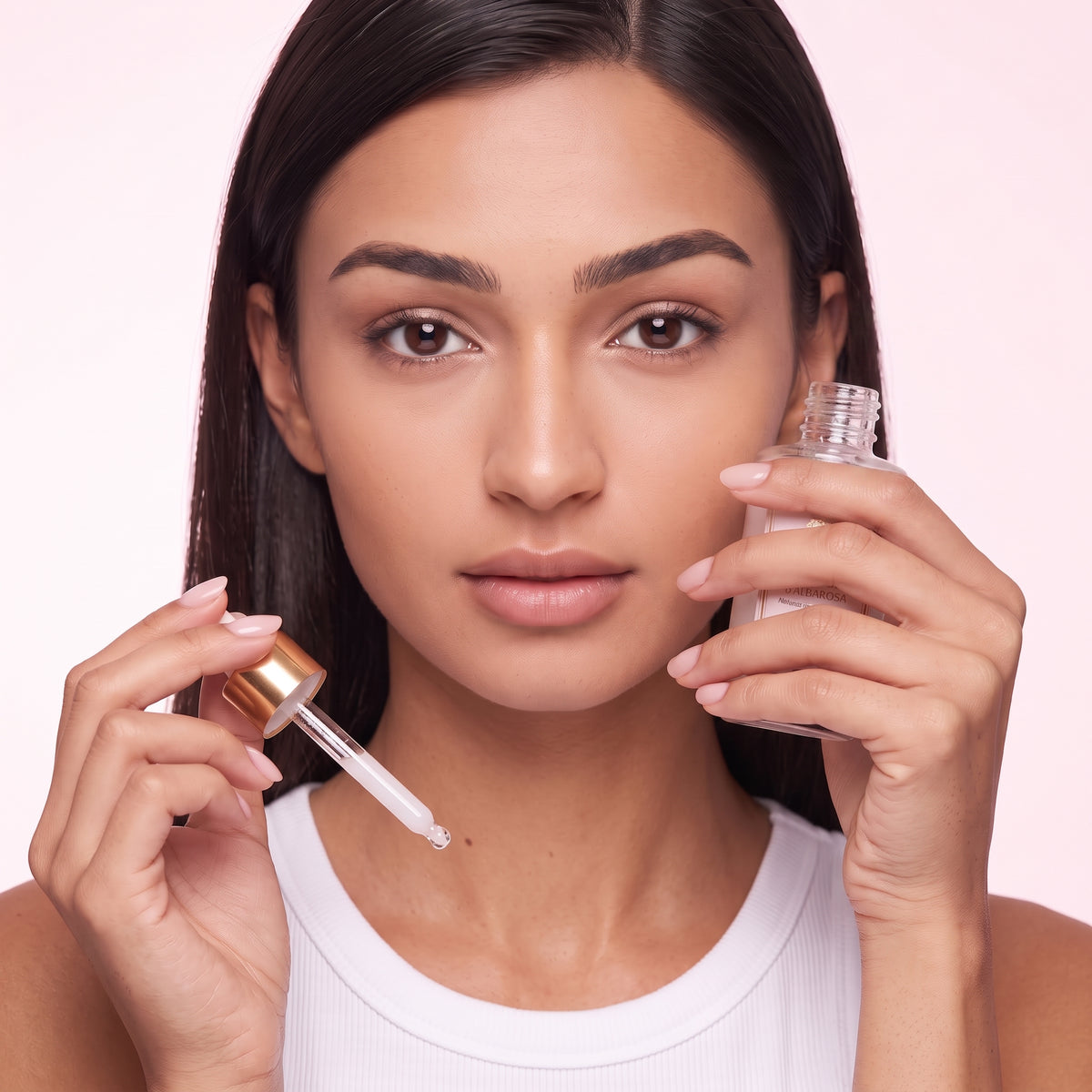 A close-up of a woman holding a clear skincare dropper bottle and its dropper, with a small clear bottle in the other hand, against a soft light pink background.