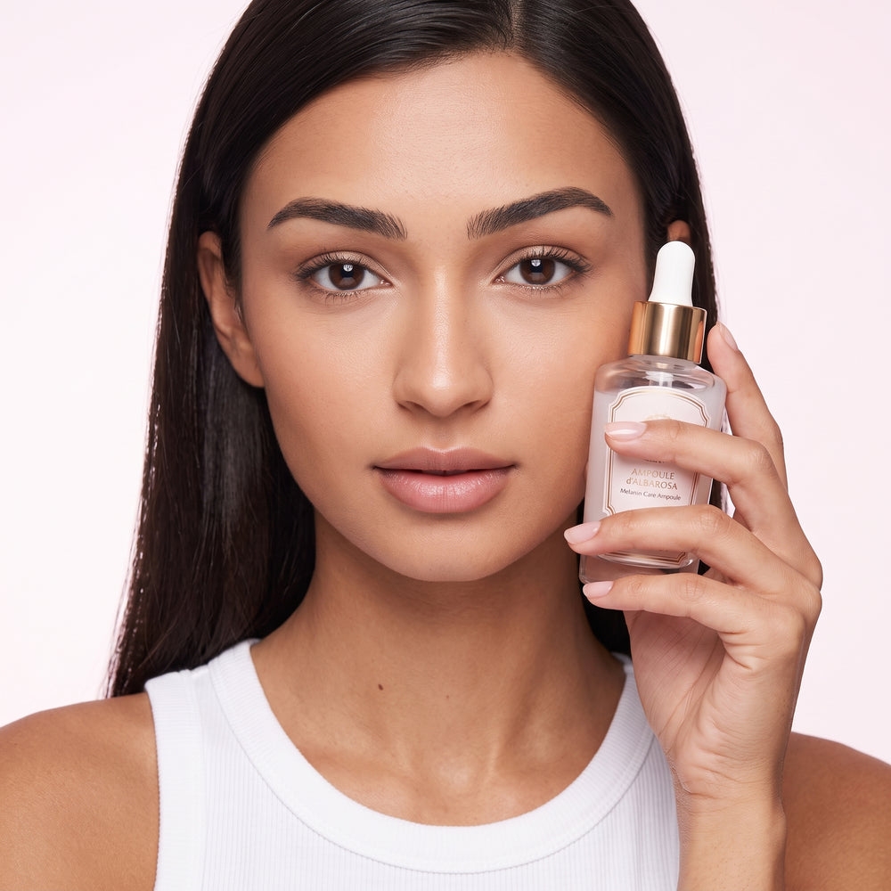 A woman with long dark hair holds a clear glass dropper bottle of Rosewater Balmy Rosewater Cleanser next to her face against a soft pink background.