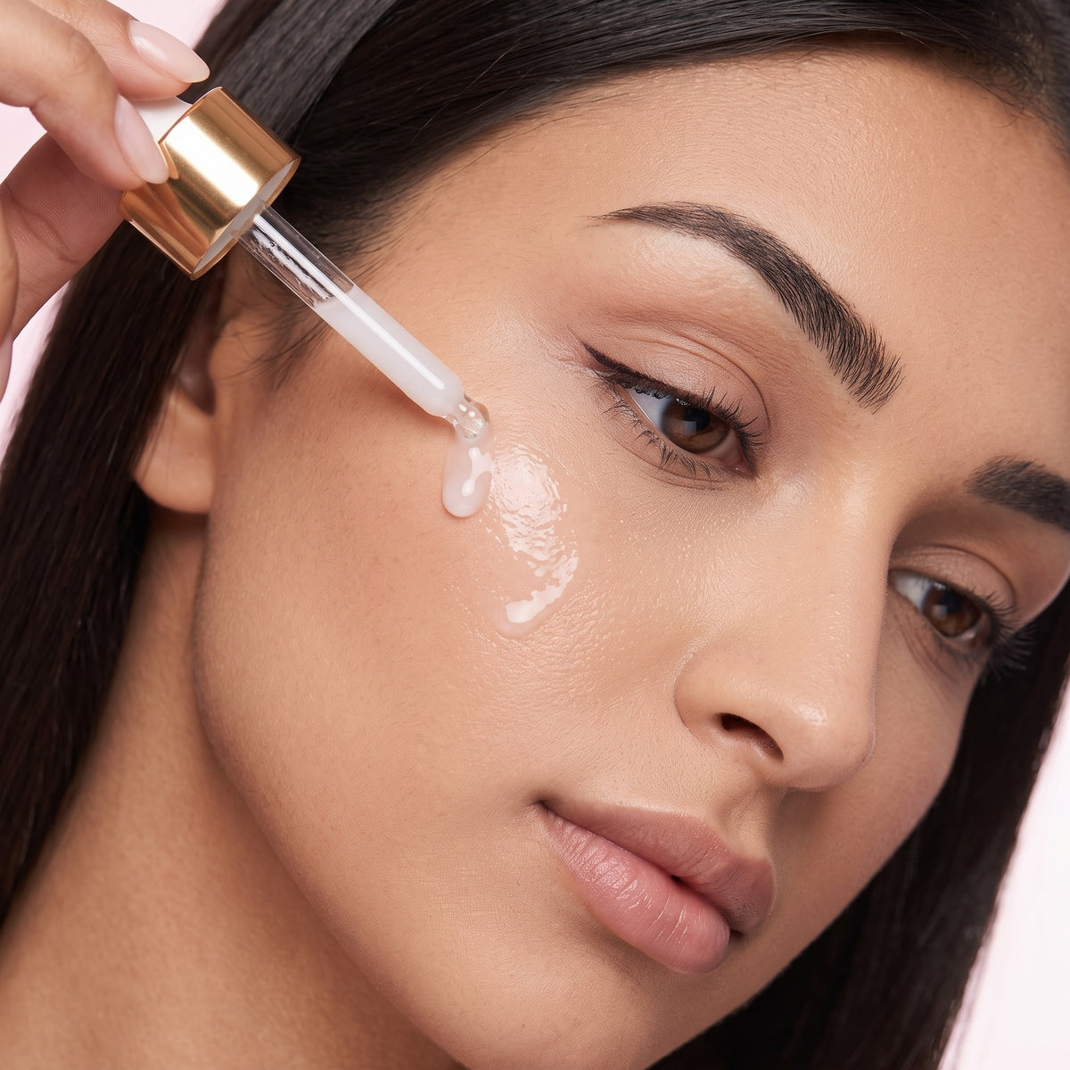 Close-up of a woman applying a clear skincare serum from a gold-capped dropper bottle onto her cheek, with a glossy droplet visible on her skin against a soft pink background.